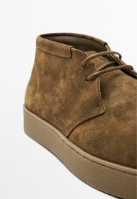 Bottines chukka en suède brun avec une surface texturée, un bout rond et des lacets bruns assortis sur une semelle en caoutchouc beige. Détails de couture minimalistes.