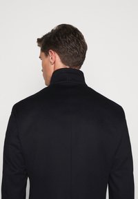 Cappotto in lana navy con colletto alto e vestibilità su misura. Presenta branding minimale sul colletto. Texture liscia e linee pulite.