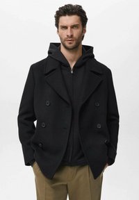 Cappotto in lana nero doppiopetto con ampi revers, abbinato a una felpa nera con zip, indossato sopra pantaloni beige. Texture liscia, senza modelli visibili.
