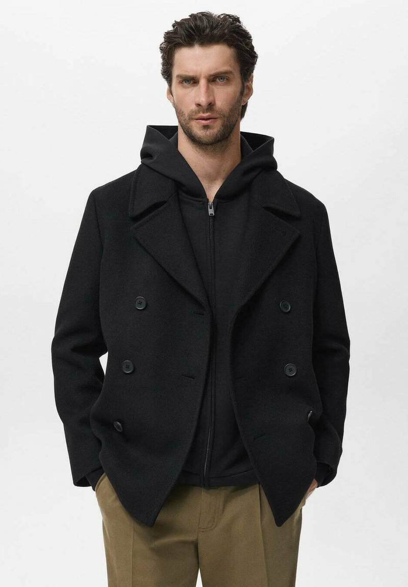 Cappotto in lana nero doppiopetto con ampi revers, abbinato a una felpa nera con zip, indossato sopra pantaloni beige. Texture liscia, senza modelli visibili.