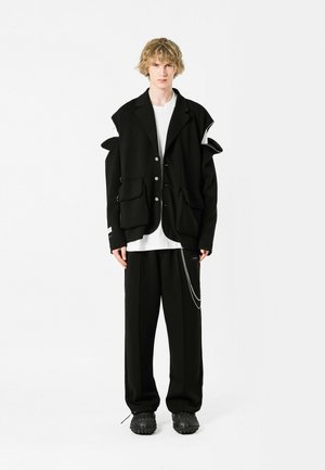 Chaqueta oversize negra con acentos distintivos en los hombros, cinco botones, grandes bolsillos, combinada con pantalones negros y calzado texturizado.