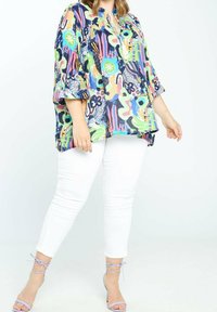 Kvinde iført bluse med multicolor abstrakt print, hvide skinny bukser og lyse lilla stroppede hæle, der står på en hvid baggrund.
