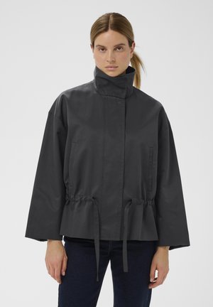 Sötétszürke, rövidített dzseki magas gallérral, rugalmas derékpánttal és oldalzsebekkel. Simább textúrájú, bő szabású, húzózsinóros részletekkel.