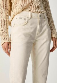 Jean en denim beige clair avec une coupe droite, présentant des poches avant, des accessoires en métal argenté et des coutures contrastantes. Porté avec un haut en tricot texturé.