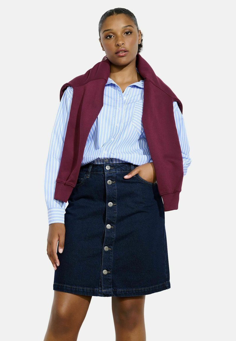 Denim krilo, temno modro, do kolen z zapiranjem spredaj na gumb; kombinirano s svetlo modro in belo prugasto srajco ter maroon puloverjem.