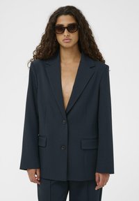 Sötét tengerészkék zakó strukturált szövetből, bevágott hajtókákkal, kétgombos zárral és két első zsebbel.