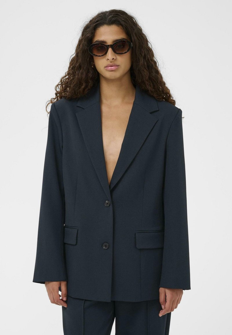 Sötét tengerészkék zakó strukturált szövetből, bevágott hajtókákkal, kétgombos zárral és két első zsebbel.