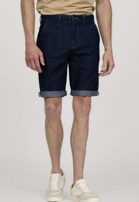 Shorts en denim bleu foncé avec des revers, arborant un design classique et des poches avant. Portés avec des baskets blanches.