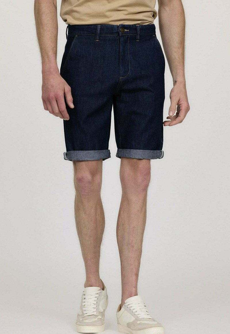 Shorts en denim bleu foncé avec des revers, arborant un design classique et des poches avant. Portés avec des baskets blanches.