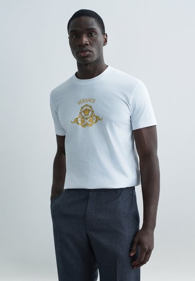 T-shirt bianca in cotone con logo Versace in oro a rilievo e design elaborato, abbinata a pantaloni grigio scuro dalla trama strutturata.
