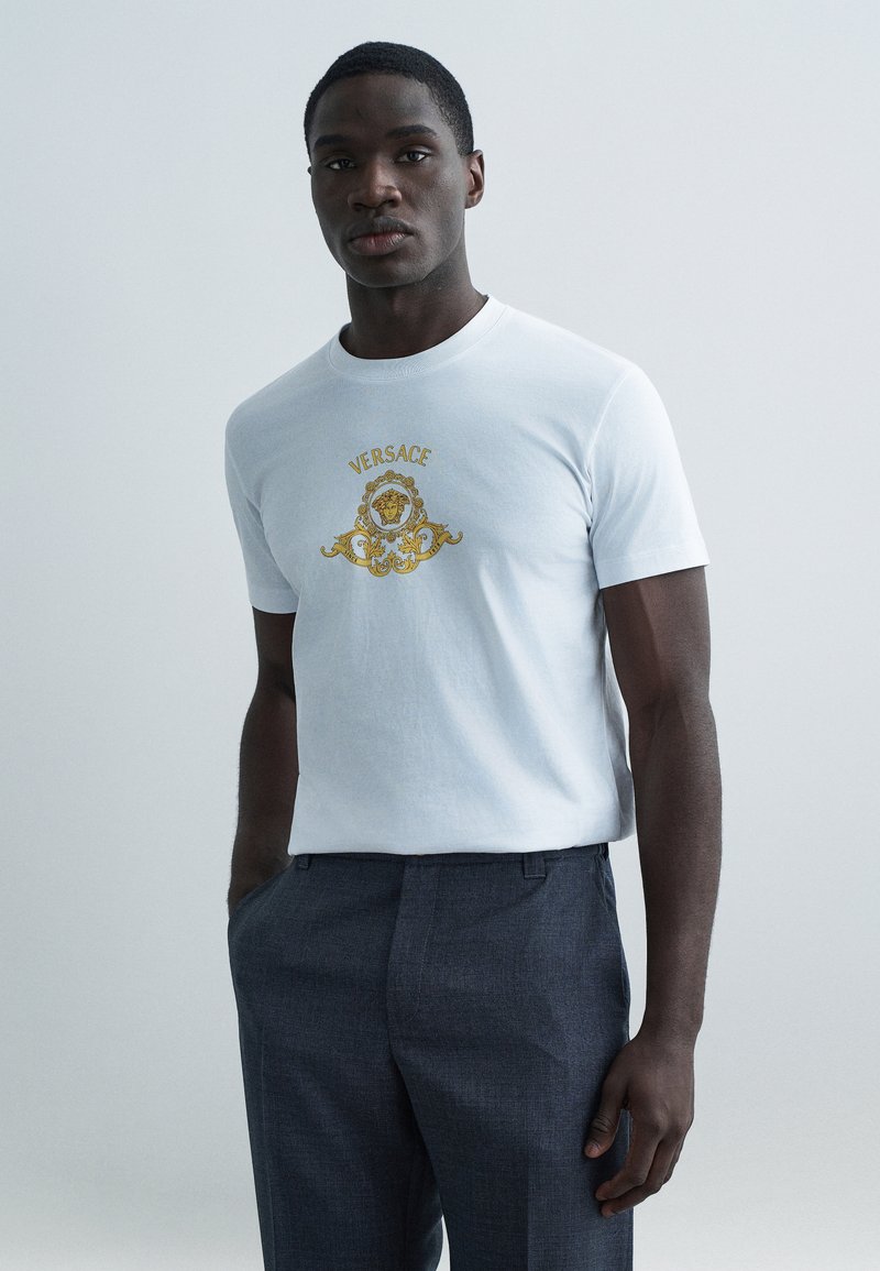 Hvid bomulds t-shirt med guldpræget Versace-logo og indviklet mønster, kombineret med mørkegrå, teksturerede bukser.