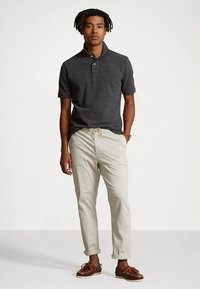 Σκούρο γκρι πόλο μπλουζάκι με κοντά μανίκια και μικρό πράσινο λογότυπο, σε συνδυασμό με ανοιχτό μπεζ παντελόνι και καφέ παπούτσια τύπου boat shoes.