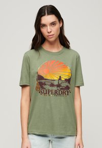 Camiseta de algodón verde oliva con un estampado gráfico que presenta un sol, montañas y el texto "SUPERDRY" en colores marrón y naranja. Manga corta, cuello redondo.