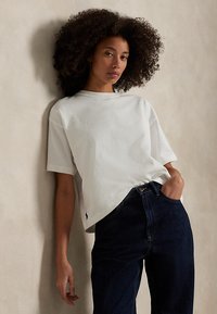Vrouw met krullend haar die tegen een beige muur leunt, draagt een wit T-shirt met korte mouwen en donkerblauwe spijkerbroek met één hand in de zak.