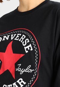Schwarzes Shirt mit einem großen roten Stern und weißem "Converse All Star"-Text mit der Unterschrift "Taylor", die den Stern auf der Vorderseite umgibt.