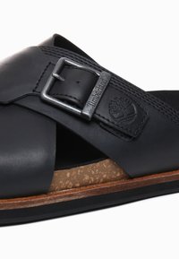 Sandale en cuir noir avec une large sangle, dotée d'une boucle en métal et d'un coussin en liège. Surface texturée et détail de semelle en bois.