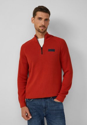 Roter, strukturierten Pullover mit halbem Reißverschluss, mit einem navyfarbenen Etikett auf der Brust und gerippten Bündchen, getragen über Jeans.