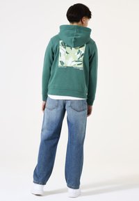 Felpa verde con un design grafico di piante sul retro, abbinata a jeans azzurri chiari. La felpa presenta un cappuccio con cordino e polsini a coste.