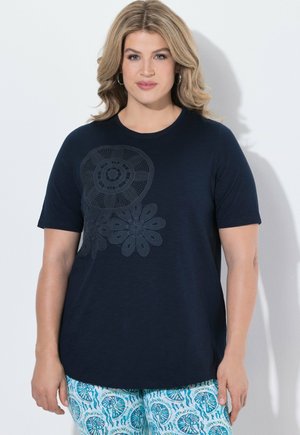 Kobieta o blond włosach do ramion, ubrana w granatową bluzkę z krótkim rękawem z kwiatową koronką oraz niebieskie spodnie we wzory.