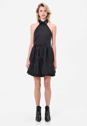 Donna che indossa un vestito nero senza maniche con scollo a halter, gonna svasata e stivaletti neri, in piedi davanti a uno sfondo bianco semplice.