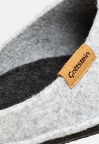Szare filcowe kapcie z czarną podeszwą i brązową skórzaną metką z wytłoczonym napisem "Gottstein". Gładka faktura, okrągły kształt.
