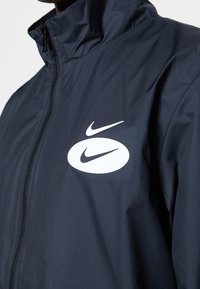 Zwarte jack met een voorsluiting en twee witte Nike swoosh-logo's op de linkerborst, waarvan de ene in een ovale vorm boven de andere is geplaatst.