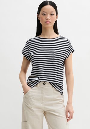 Vrouw met lang, steil haar draagt een zwart-wit gestreept topje met korte mouwen, ingestopt in beige broek met hoge taille en zakken, staand tegen een witte achtergrond.