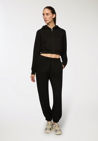 Donna in piedi che indossa un hoodie nero corto con zip, pantaloni jogger neri coordinati e sneakers chunky beige, mentre guarda di lato.