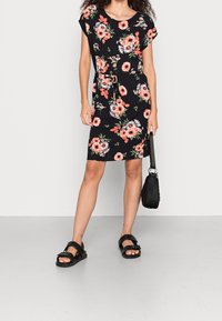 Blumenkleid mit schwarzem Grund und pinken sowie weißen Blumen, kurzen Ärmeln, einem Kordelzug in der Taille und einem knielangen Schnitt. Schwarze Handtasche und Sandalen.