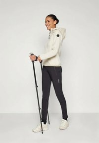 Heled värvi zipp-up kapuutsiga jakki mustade detailidega, koos sobivate mustade pükste ja valgete spordijalatsitega, hoides matkakeppe.