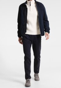 Marint bombarjacka, vit ribbad tröja, mörka byxor, grå mockaskor; visad modell i en gångposition mot en ljus bakgrund.