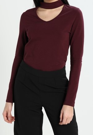 Kvinde iført en bordeauxrød top med lange ærmer og chokerhalsudskæring samt sorte bukser med høj talje mod en ensfarvet baggrund.