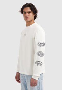 Wit T-shirt met lange mouwen en een ronde hals. Voorzien van zwarte bedrukte logo's op beide mouwen. Gemaakt van een zachte, lichte stof. Regular fit.