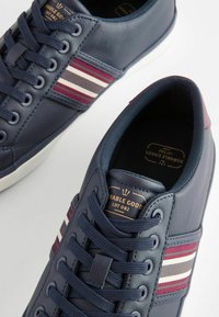 Kék szintetikus bőr sneaker, kontrasztos bordó és krém csíkokkal, kerek orral és fűzős záródással. Textúrált bélés és márkajelzés.
