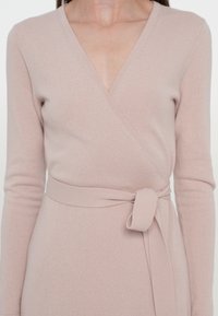 Femme portant une robe portefeuille beige clair à manches longues, avec un col en V et une ceinture en tissu nouée à la taille.