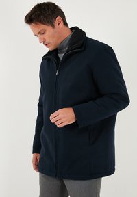 Vīrietis valkā tumši zilu zipa mēteli pār pelēku turtleneck kreklu, skatās uz leju, ar vienu roku tuvu rāvējslēdzējam.