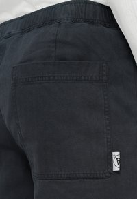 Close-up of dark gray herringbone patterned pants with back patch pocket and small white brand tag on the right side.