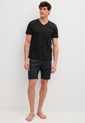 Young man standing barefoot wearing a black V-neck T-shirt with pocket and dark patterned shorts against a plain light background.