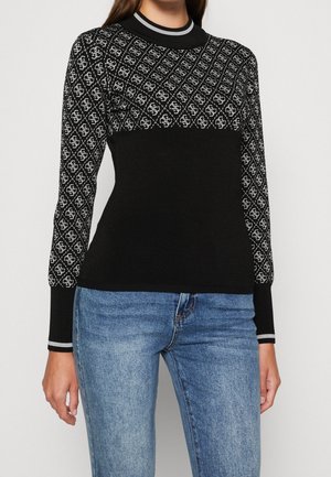 Femme portant un pull noir avec un motif losange gris sur le haut de la poitrine et les manches, associé à un jean bleu, debout devant un fond uni.