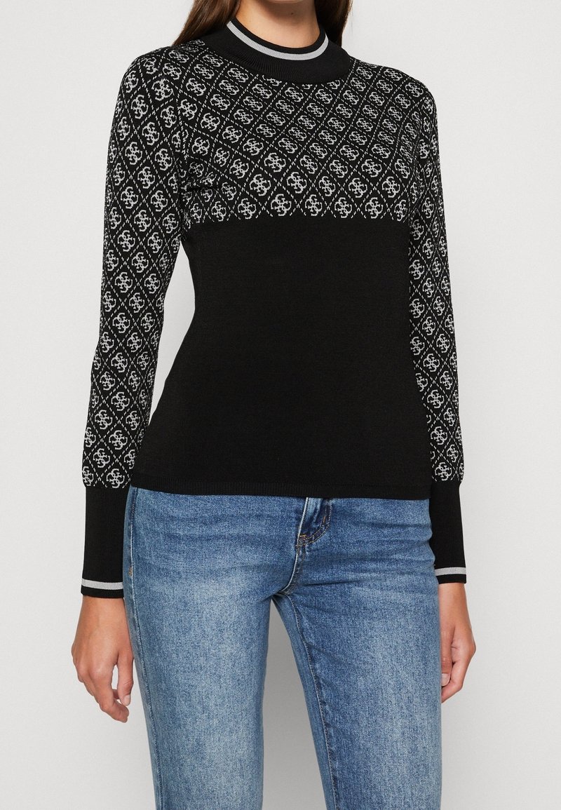 Femme portant un pull noir avec un motif losange gris sur le haut de la poitrine et les manches, associé à un jean bleu, debout devant un fond uni.