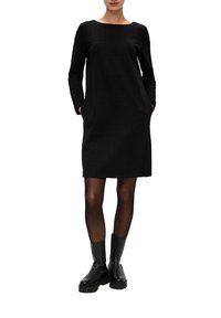 Czarna sukienka z długim rękawem, wykonana z teksturowanej tkaniny, z okrągłym dekoltem, dwiema kieszeniami i długością do kolan. Noszona z czarnymi kozakami.