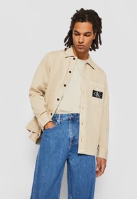 Gai beige krekls ar jaku, melnām pogām un krūšu kabatu ar logotipu, apvienots ar brīvām ziliem džinsiem.