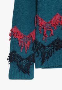 Maglione lavorato a maglia in teal con accenti frangiati in rosso e nero a contrasto in un motivo a zigzag lungo i bordi inferiori. Texture morbida.