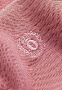 Tela rosa con un tejido texturizado, que presenta un logotipo blanco bordado rodeado de hojas de laurel y un pequeño detalle de corona.