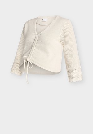 Cardigan blanc en maille avec col en V, coupe courte, manches 3/4 agrémentées de détails en dentelle, et fermeture à nouer sur le devant.