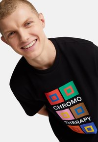 Camiseta de algodón negra con un diseño gráfico rectangular colorido que presenta el texto "TERAPIA DE COLOR" en varios colores. Cuello redondo estándar.