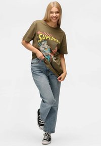 Camiseta de algodón verde oliva con un gráfico de "Superman", combinada con unos vaqueros de color azul claro y unas zapatillas en blanco y negro.