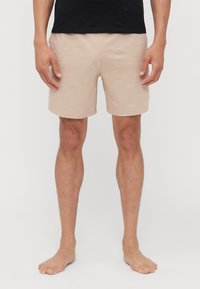 Beige badeshorts med et teksturert logomønster, elastisk midje og en midt-lår lengde, stylet med en svart skjorte og bare føtter.