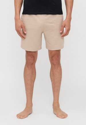 Beige Badehose mit strukturiertem Logo-Muster, elastischem Bund und einem Schnitt bis zur Mitte des Oberschenkels, kombiniert mit einem schwarzen Shirt und barfuß.