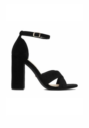 Czarna sandał na wysokim obcasie zamszowy z masywnym obcasem, paskiem wokół kostki z klamrą oraz skrzyżowanymi paskami na palcach.